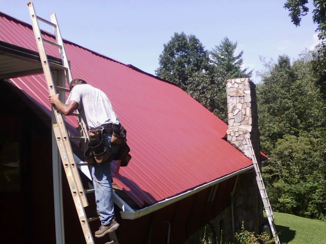 Roofer on a ladder beside a red metal roof and stone chimney — Soffit & Fascia Repair by Roofing Brothers in Mission Hills